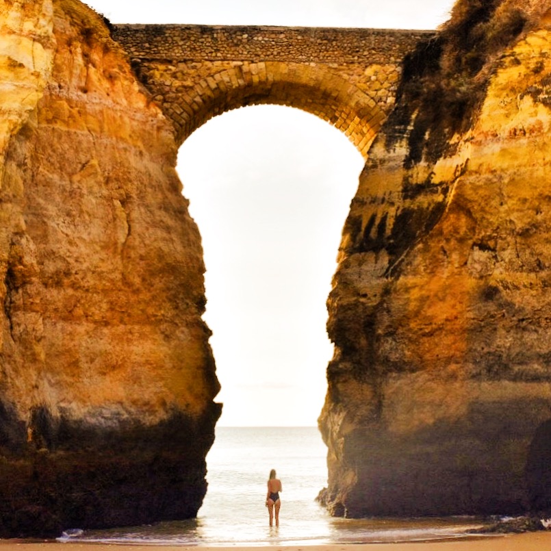 Sarah Latham standing on the beach at the ocean underneath bridge arch between two cliffs in Lagos, Portugal