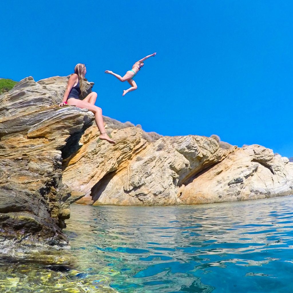 One girl jumping off a cliff into the ocean while another girl watches from the side line in Ios, Greece