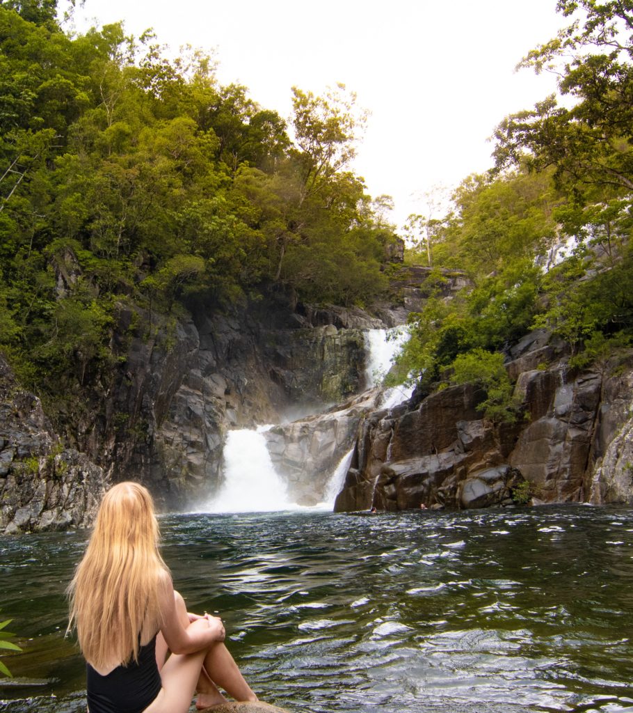 Girl sitting with back to camera overlooking large waterfall and waterhole