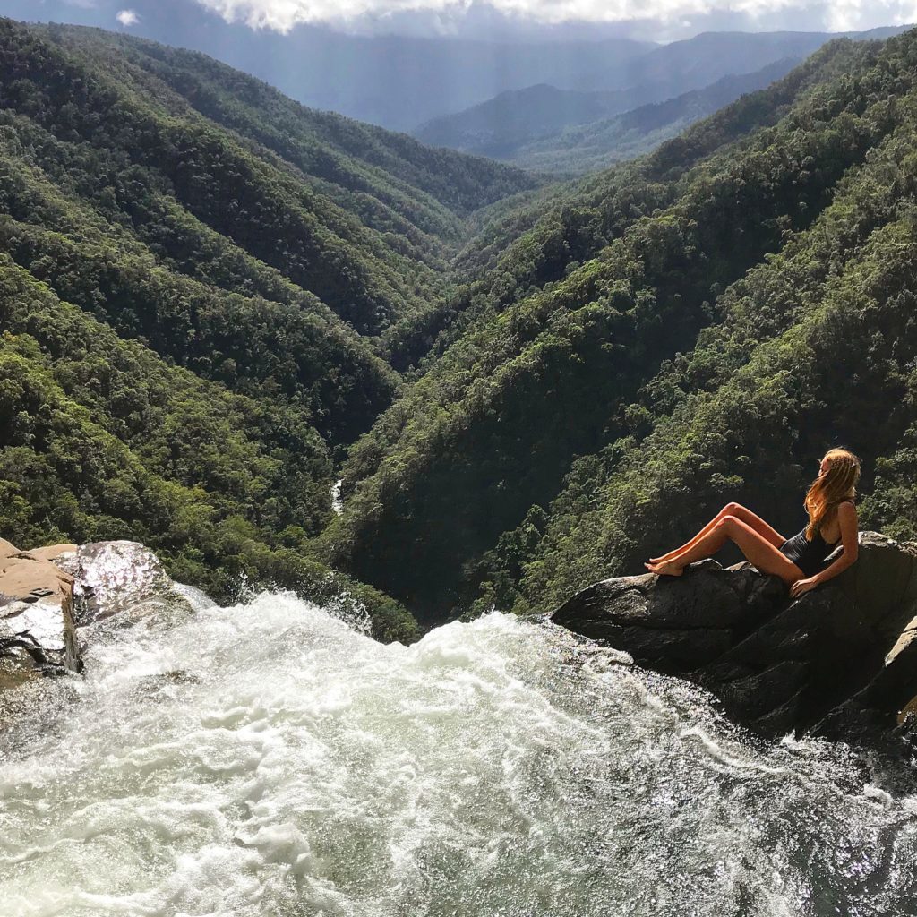 Girl sitting on edge of top of Windin Falls overlooking valley below