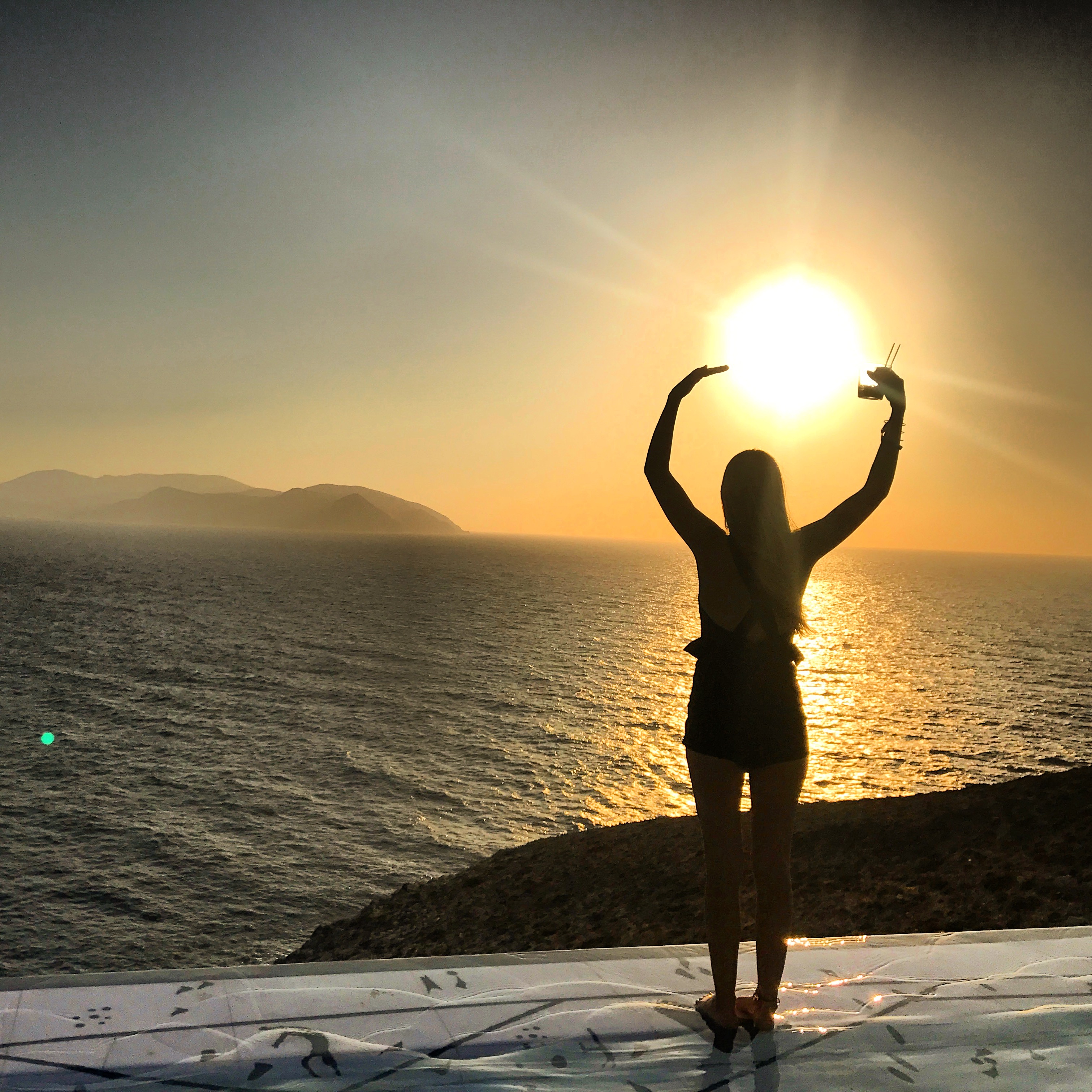 Girl sillohouetted against the sunset standing on top of an infinity pool in Ios, Greece