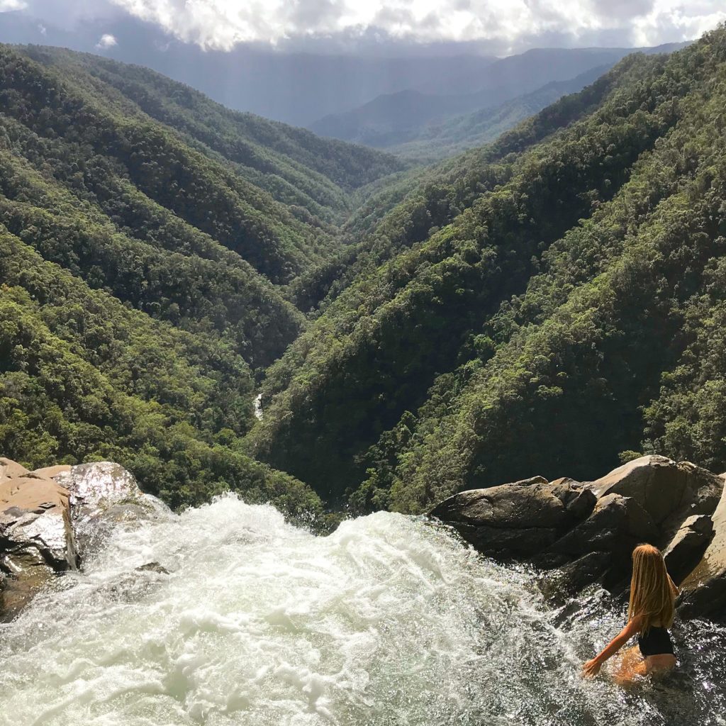 girl sitting on the edge of the top of Windin Falls waterfall looking out over valley